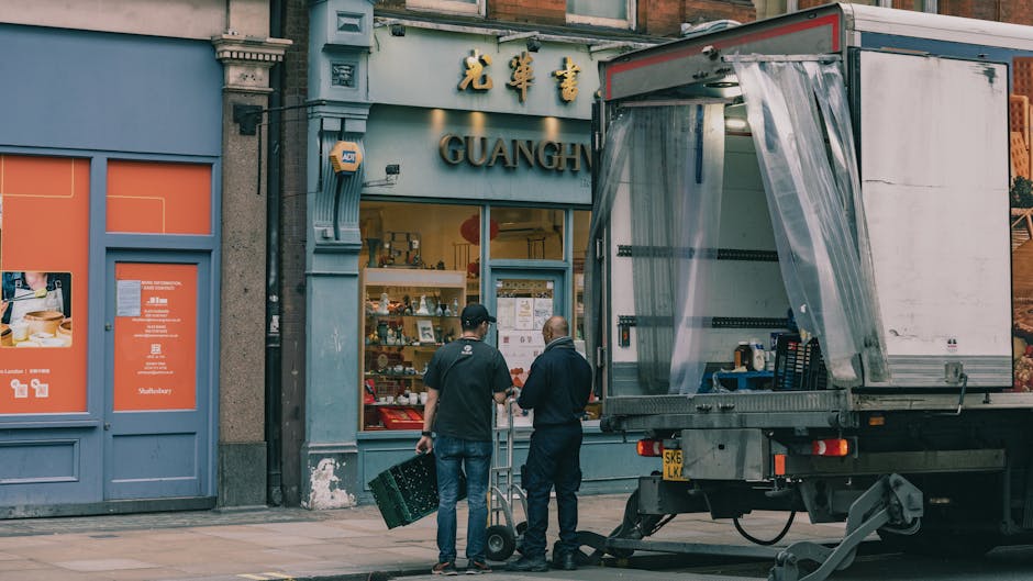 Two men stand outside a storefront with a blue façade, marked with gold Chinese characters and the word 'GUANG' above the entrance, supporting a home relocation or furniture transport process. One man, dressed in a dark jacket and jeans, holds a black plastic crate, while the other, in a dark jacket and dark pants, uses a hand truck to move packed boxes. In front of them, a large, open-sided removal van with a transparent plastic curtain on the side is parked on the pavement, loaded with wrapped furniture, cardboard boxes, and packing materials. The scene is set during daylight, with other shops visible nearby. This visual depicts professional packing and loading activities typical of house removals services, as offered by South Kensington Removals, emphasizing organized furniture transport and packing during a house move or relocation process.