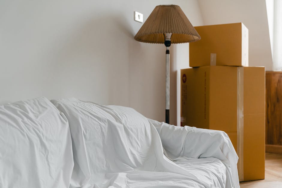 Inside a room during a home relocation process, there are four cardboard boxes stacked in the corner near a wooden sideboard; the boxes are sealed with packing tape and vary in size, with the largest at the bottom. To the left, a white sofa draped with a protective fabric cover is positioned against a plain, light-colored wall. On the wall above the sofa, there is a white light switch. A floor lamp with a beige pleated lampshade and a silver metallic stand stands next to the boxes, providing soft lighting to the area. Wooden flooring is visible beneath the furniture and boxes. The scene captures a moment of packing and moving, with packages ready for transportation, and is indicative of the detailed process involved in house removals offered by South Kensington Removals, emphasizing careful packing, furniture transport, and loading procedures typical in professional relocation services.
