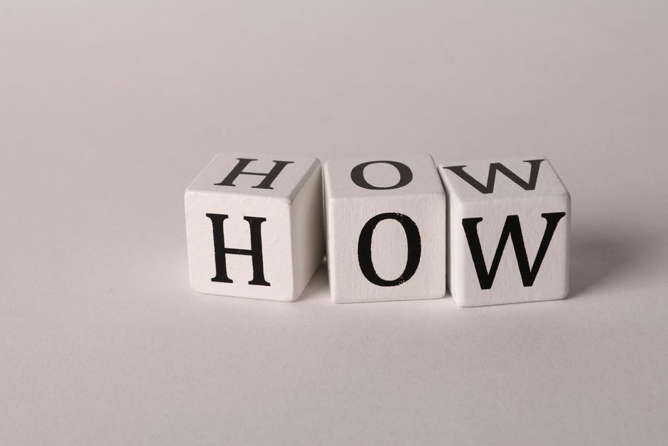 Close-up image of three white wooden blocks arranged in a row on a light grey surface, each displaying a black letter that together spell 'HOW'. The blocks are positioned horizontally in the foreground, with the first block slightly tilted, indicating movement or handling. This visual relates to house removals and moving logistics, highlighting the process of packing and preparing items for a smooth home relocation. The neutral background and lighting emphasize the simplicity and clarity of the scene, reflecting the professional organization involved in furniture transport, packing, and loading procedures. South Kensington Removals, a company offering removals services, may use such imagery to illustrate the initial planning and careful packaging stages of a comprehensive moving process advertised on their website, demonstrating attention to detail during a typical house move.