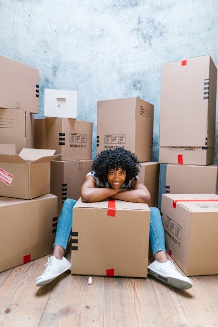 A young woman with curly black hair, wearing a white t-shirt, jeans, and white sneakers, sitting on a light wooden floor amid numerous cardboard boxes of varying sizes, some sealed with red or black tape and others open with packing materials visible. She is smiling and resting her arms on a closed box in front of her, surrounded by stacked boxes inside a room with a light blue textured wall. The scene illustrates a home relocation process with a focus on packing activities, supported by South Kensington Removals’ services, and depicts the logistics involved in furniture transport and the moving process.