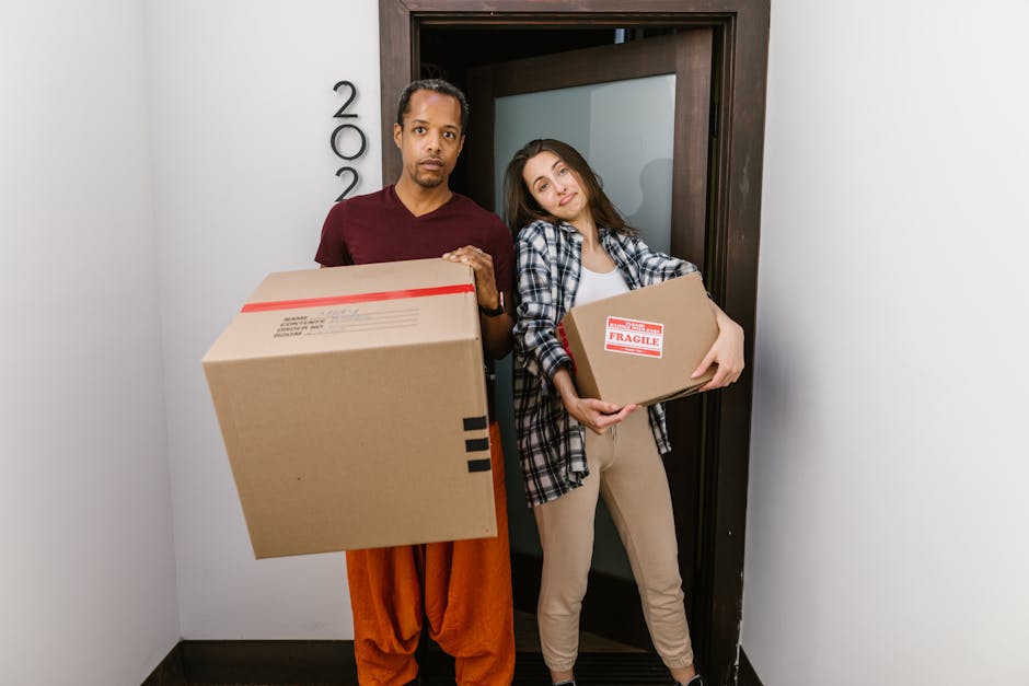 A man and a woman stand inside a building near an open doorway, each holding cardboard boxes as part of a home relocation process. The man, dressed in a burgundy T-shirt and orange trousers, holds a large box labeled with a barcode. The woman, wearing a plaid shirt over a white top and beige trousers, holds a smaller box marked with a red 