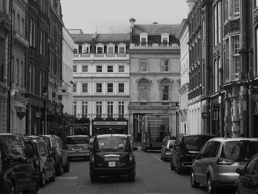 Black and white photograph of a narrow urban street in South Kensington, showing multiple parked cars lining each side of the road in front of elegant multi-storey buildings with detailed facades and 
