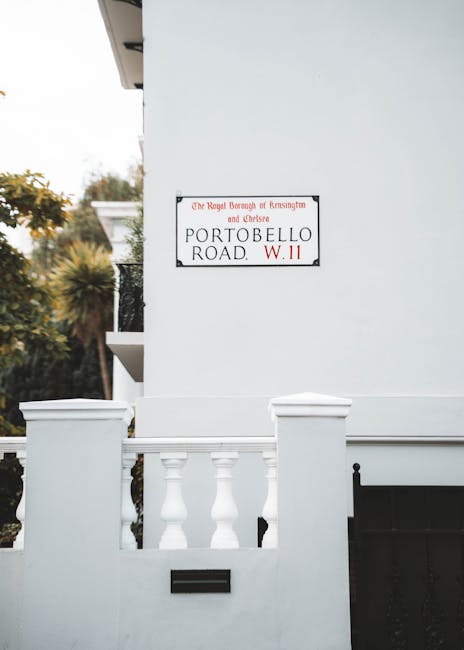 A white exterior wall of a residential property displaying a street sign reading 'Portobello Road, W.11' beneath a black metal mailbox. The property features a short white wall with decorative balustrades, and a small section of a black gate is visible to the right. Inside the property, partially seen behind the white wall, are several cardboard boxes and protective packing materials such as bubble wrap and plastic wrapping, likely prepared for a home relocation or furniture transport. The background shows some greenery, including trees and bushes, indicating an outdoor setting in Kensington and Chelsea. The scene suggests an ongoing packing or moving process, with South Kensington Removals providing professional assistance with house removals and furniture transport services, aligned with local council regulations.