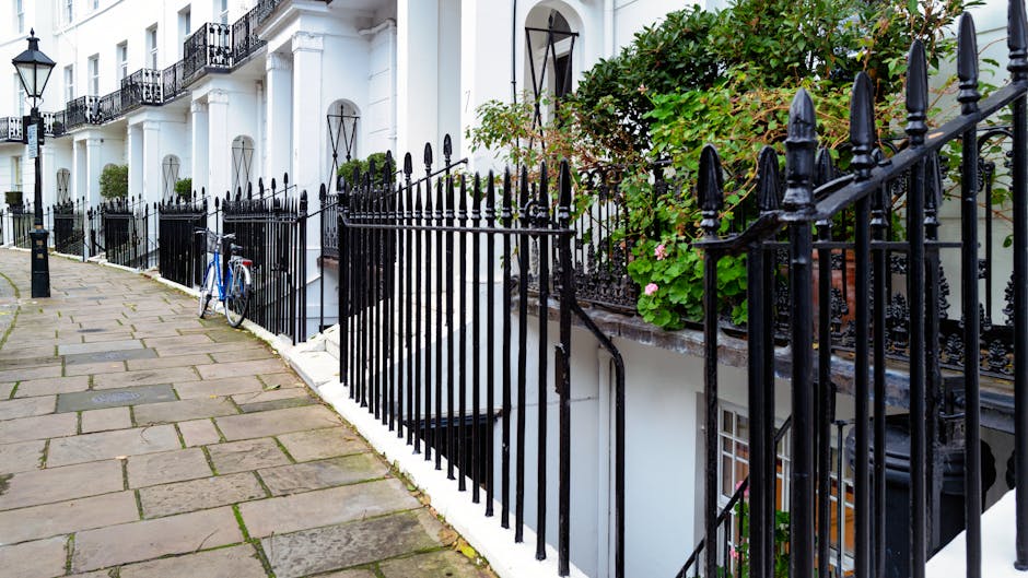 A row of white terraced houses with black wrought iron railings along the pavement in Kensington and Chelsea, London. The railings surround small front gardens with green plants and flowers, some supported by brick walls. An outdoor blue bicycle is parked against one of the railings near a doorway, which leads to a basement entrance partially visible below street level with a black metal railing steps. The sidewalk is paved with rectangular stone slabs, and the scene is illuminated by natural daylight, with some foliage visible on the right side of the image. This setting depicts an urban residential area suitable for house removals or furniture transport, where careful handling of items and adherence to local council permit rules, as outlined by South Kensington Removals, might be required during home relocation efforts.
