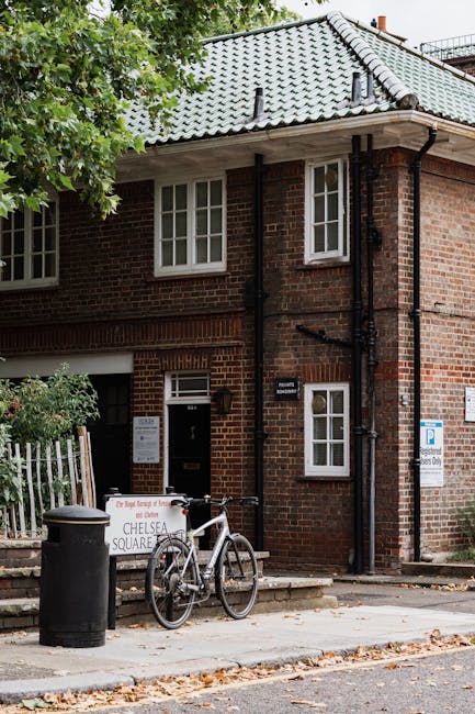 A white exterior wall of a residential property displaying a street sign reading 'Portobello Road, W.11' beneath a black metal mailbox. The property features a short white wall with decorative balustrades, and a small section of a black gate is visible to the right. Inside the property, partially seen behind the white wall, are several cardboard boxes and protective packing materials such as bubble wrap and plastic wrapping, likely prepared for a home relocation or furniture transport. The background shows some greenery, including trees and bushes, indicating an outdoor setting in Kensington and Chelsea. The scene suggests an ongoing packing or moving process, with South Kensington Removals providing professional assistance with house removals and furniture transport services, aligned with local council regulations.