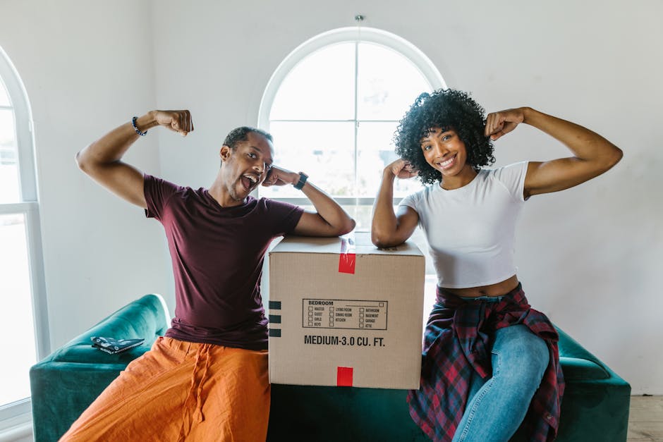 A man and woman, both smiling and flexing their arm muscles, pose inside a bright room with large arched windows, standing next to a cardboard box labeled for a bedroom with measurements of medium size, indicating a house move or packing activity. The man, wearing a maroon T-shirt and orange pants, is sitting on a teal sofa, while the woman, dressed in a white cropped T-shirt and jeans with a red and black plaid shirt tied around her waist, is kneeling beside the sofa. The scene suggests a casual moment during home relocation, with the cardboard box prepared for furniture transport and packing, and a focus on the early stages of a house removals process. The room features natural daylight illuminating the space, emphasizing the context of moving and packing activities as part of an organized house removal service by South Kensington Removals.