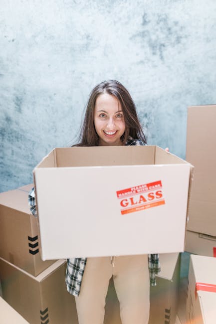 A young woman with curly black hair, smiling, sitting on the floor inside a room surrounded by numerous cardboard moving boxes of various sizes, some with labels and red packing tape. She is resting her arms on a medium-sized box in front of her, wearing casual clothing and white sneakers. The room has a light blue textured wall and a wooden floor, indicating an ongoing home relocation process. This scene illustrates packing and moving activities, supported by South Kensington Removals for house relocation services within the UK.