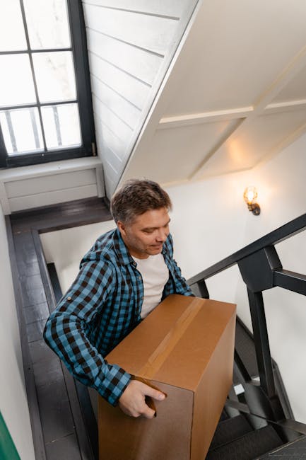A man with short brown hair, wearing a blue and black checkered shirt over a white t-shirt, is carrying a cardboard box up a narrow staircase inside a residential property. The staircase has dark wooden steps and black metal railings, with a white wall on one side and a window with black framing next to it, allowing natural light into the space. An interior wall with a wall-mounted light fixture is visible, highlighting the confined safe passage for home relocation or furniture transport activities, as handled by South Kensington Removals. The overall environment suggests a typical staircase in a home, presenting challenges for moving larger items like furniture or boxes along a tight, steep, and confined stairway.
