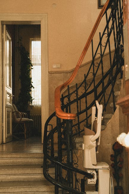 A narrow staircase inside a property with a black wrought iron balustrade, a wooden handrail, and beige marble steps. At the bottom of the stairs, a white portable stair lift is partially visible, positioned against the wall to assist with home mobility. In the background, a doorway reveals a room with natural light coming through a large window, showing a wooden bench with a cushion and a tall plant beside the window. The surrounding walls are finished with a neutral-toned wallpaper, and a light switch is mounted on the wall beside the staircase. The environment appears to be prepared for a furniture transport or home relocation process, with the staircase presenting limited space for moving large items. South Kensington Removals is experienced in navigating such challenging staircases during house moving and furniture transport services, emphasizing careful packing, loading, and maneuvering of household items in confined environments.