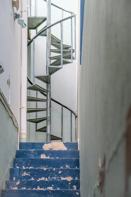 A man with short brown hair, wearing a blue and black checkered shirt over a white t-shirt, is carrying a cardboard box up a narrow staircase inside a residential property. The staircase has dark wooden steps and black metal railings, with a white wall on one side and a window with black framing next to it, allowing natural light into the space. An interior wall with a wall-mounted light fixture is visible, highlighting the confined safe passage for home relocation or furniture transport activities, as handled by South Kensington Removals. The overall environment suggests a typical staircase in a home, presenting challenges for moving larger items like furniture or boxes along a tight, steep, and confined stairway.
