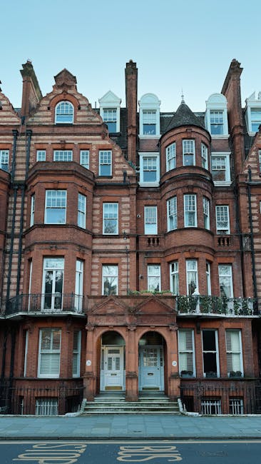 Photograph of a multi-storey Victorian residential building with a red brick facade, decorative arch windows, and distinctive stone detailing characteristic of homes along Old Brompton Road in South Kensington. The building features several bay windows, small balconies with wrought iron railings on the lower floors, and a central entrance with a set of steps leading up to double doors. Above the entrance, there are arched window openings and several chimneys protrude from the roofline. The image is taken from the street, showing the building front and pavement below, with clear daylight and a blue sky in the background. This type of architecture is typical for area house removals and residential relocations, highlighting the importance of professional moving services like those offered by South Kensington Removals for home relocation and furniture transport in South Kensington.