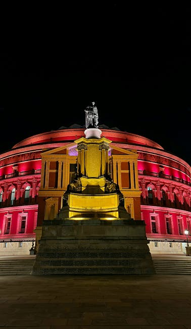 The image shows the Royal Albert Hall illuminated at night, with vibrant red and purple lighting highlighting its domed roof and classical architectural features. In the foreground, there is a stone statue situated on a raised platform, depicting a seated figure holding an object, with steps leading up to it. The setting appears to be outdoors on a paved area, with the Hall’s ornate façade and decorative elements clearly visible. The lighting emphasizes the grandeur of the historic building, which is often a key location in South Kensington, where house removals and furniture transport services are frequently required, such as those provided by South Kensington Removals. This scene captures the process of city-based relocating or delivery activities, where careful loading and unloading near notable landmarks are common aspects of home relocation or moving logistics.