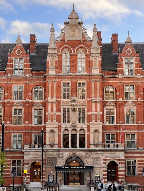 The exterior façade of the Royal College of Music building, featuring ornate red brick and stone architectural details with multiple windows and decorative turrets, situated on a street with pedestrian access. In the foreground, two individuals are seen walking on the pavement near a black entrance canopy that reads 'Royal College of Music.' The street scene includes lampposts, signage, and a few trees, with a clear sky overhead. This image relates to house removals and moving services near cultural landmarks like the Royal Albert Hall, highlighting the proximity and logistical considerations for home relocations within South Kensington, where South Kensington Removals operates.