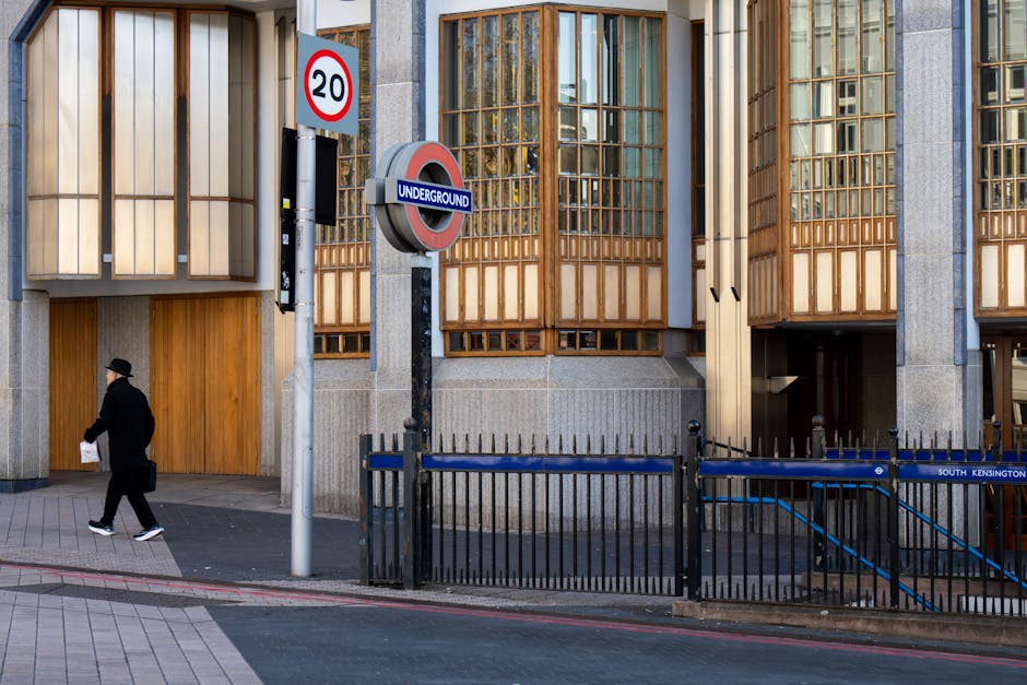 An urban street scene outside a modern building with large wooden window frames and concrete panels in South Kensington. A man dressed in black, wearing a hat, walks past a black metal fence that surrounds a staircase leading into the building. Near the entrance, there is a street sign indicating a 20-mph speed limit and an underground train station sign attached to a pole. The sidewalk is paved with rectangular concrete slabs, and a red curb runs along the street's edge. The scene appears to be during daylight with natural light illuminating the area, depicting an environment relevant for house removals and home relocation services operated by South Kensington Removals, including the process of moving furniture or packing materials outside the property.