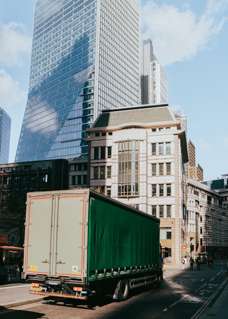 A large commercial green and beige-sided truck is parked on a city street in South Kensington, close to tall modern glass skyscrapers and historic stone buildings. The vehicle is positioned in the loading zone, with its rear doors closed, ready for furniture transport and home relocation services. The street scene includes pedestrians and cyclists in the background, indicating active urban movement. Bright daylight illuminates the scene, highlighting the contrast between the truck and the urban architecture. South Kensington Removals may use such vehicles for same-day emergency removals, coordinating loading and unloading processes efficiently in busy city environments, with careful handling of packed boxes, furniture, and appliances during the packing and moving operations.