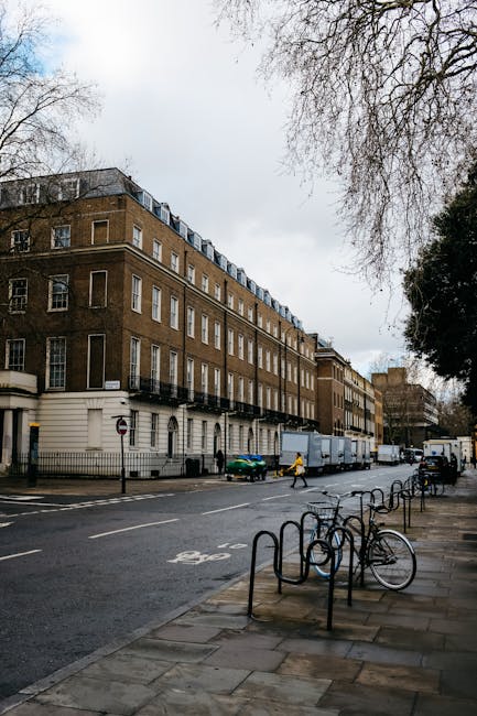 An urban street scene outside a modern building with large wooden window frames and concrete panels in South Kensington. A man dressed in black, wearing a hat, walks past a black metal fence that surrounds a staircase leading into the building. Near the entrance, there is a street sign indicating a 20-mph speed limit and an underground train station sign attached to a pole. The sidewalk is paved with rectangular concrete slabs, and a red curb runs along the street's edge. The scene appears to be during daylight with natural light illuminating the area, depicting an environment relevant for house removals and home relocation services operated by South Kensington Removals, including the process of moving furniture or packing materials outside the property.