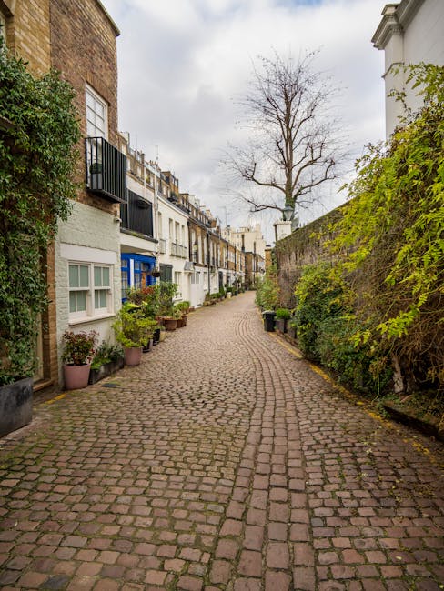 A narrow cobbled street in South Kensington lined with white and brick residential houses, some featuring small balconies and potted plants outside. A tall, leafless tree stands at the end of the street under a cloudy sky. The street appears quiet and residential, with no visible people or vehicles in motion, representing a typical area where house removals and furniture transport might begin or conclude as part of a home relocation service. South Kensington Removals' professional team could be involved in coordinating parking and logistics for moving within this historic neighbourhood, as implied by the context of the webpage about removals costs and parking tips.