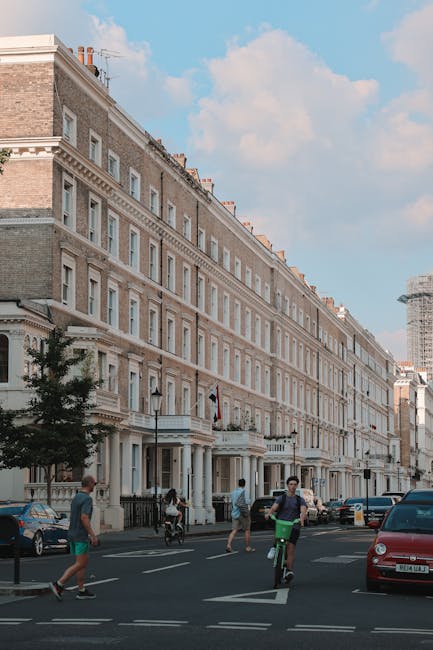 A wide view of a historic residential building in South Kensington with a classic facade, large windows, and detailed cornices, situated on a busy street with parked cars along the curb. In the foreground, pedestrians and cyclists are crossing the road; a person on a green scooter is particularly visible. The image shows fresh daylight and a partly cloudy sky indicating clear weather. Several individuals are walking near the building’s entrance, which features decorative columns and black railings, suggesting a typical home relocation or furniture transport scene. The scene portrays urban activity consistent with the kind of moving logistics managed by South Kensington Removals, including people on foot and bicycle traffic typical of London’s residential streets. The surroundings and the vehicle parking indicate a well-maintained area suitable for professional removals and packing services, aligning with targeted house removals for the SW7 postcode.