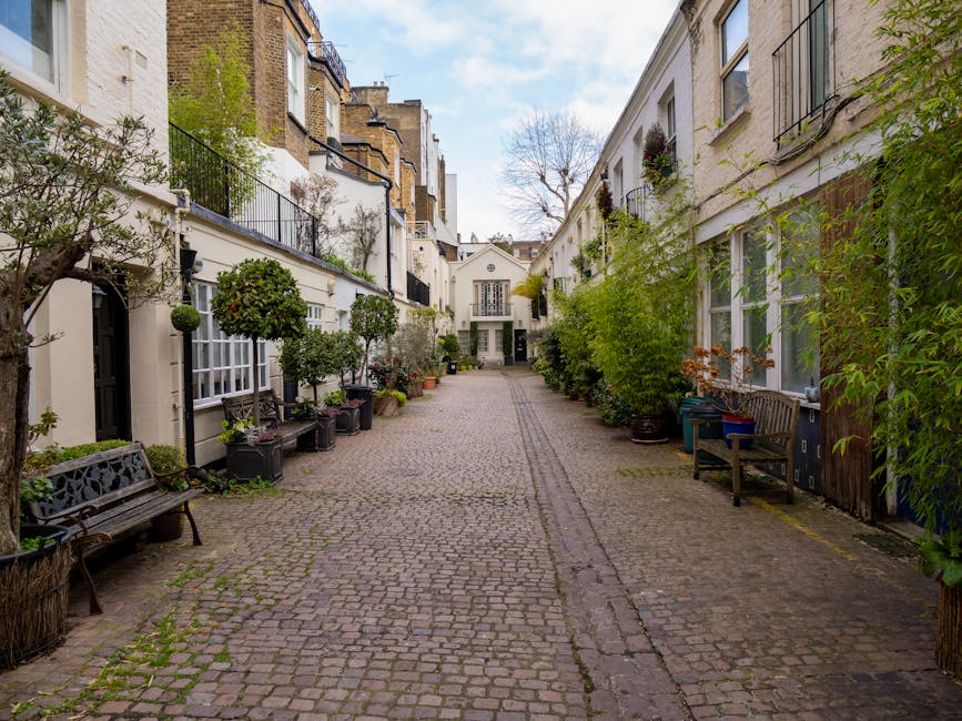 A narrow cobbled street in South Kensington lined with white and brick residential houses, some featuring small balconies and potted plants outside. A tall, leafless tree stands at the end of the street under a cloudy sky. The street appears quiet and residential, with no visible people or vehicles in motion, representing a typical area where house removals and furniture transport might begin or conclude as part of a home relocation service. South Kensington Removals' professional team could be involved in coordinating parking and logistics for moving within this historic neighbourhood, as implied by the context of the webpage about removals costs and parking tips.