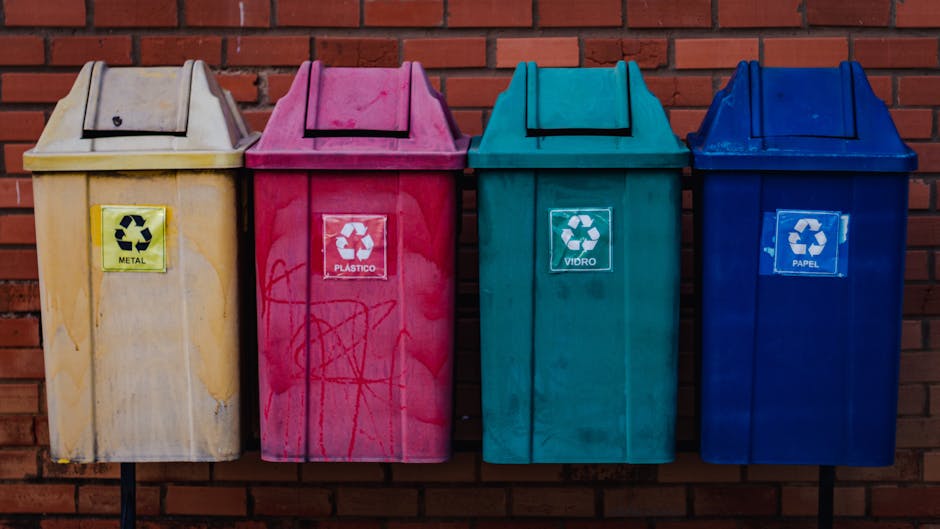 A row of four outdoor recycling bins positioned against a red brick wall, each bin painted in different colors—yellow, pink, green, and blue—clearly labeled with recyclable material categories: metal,